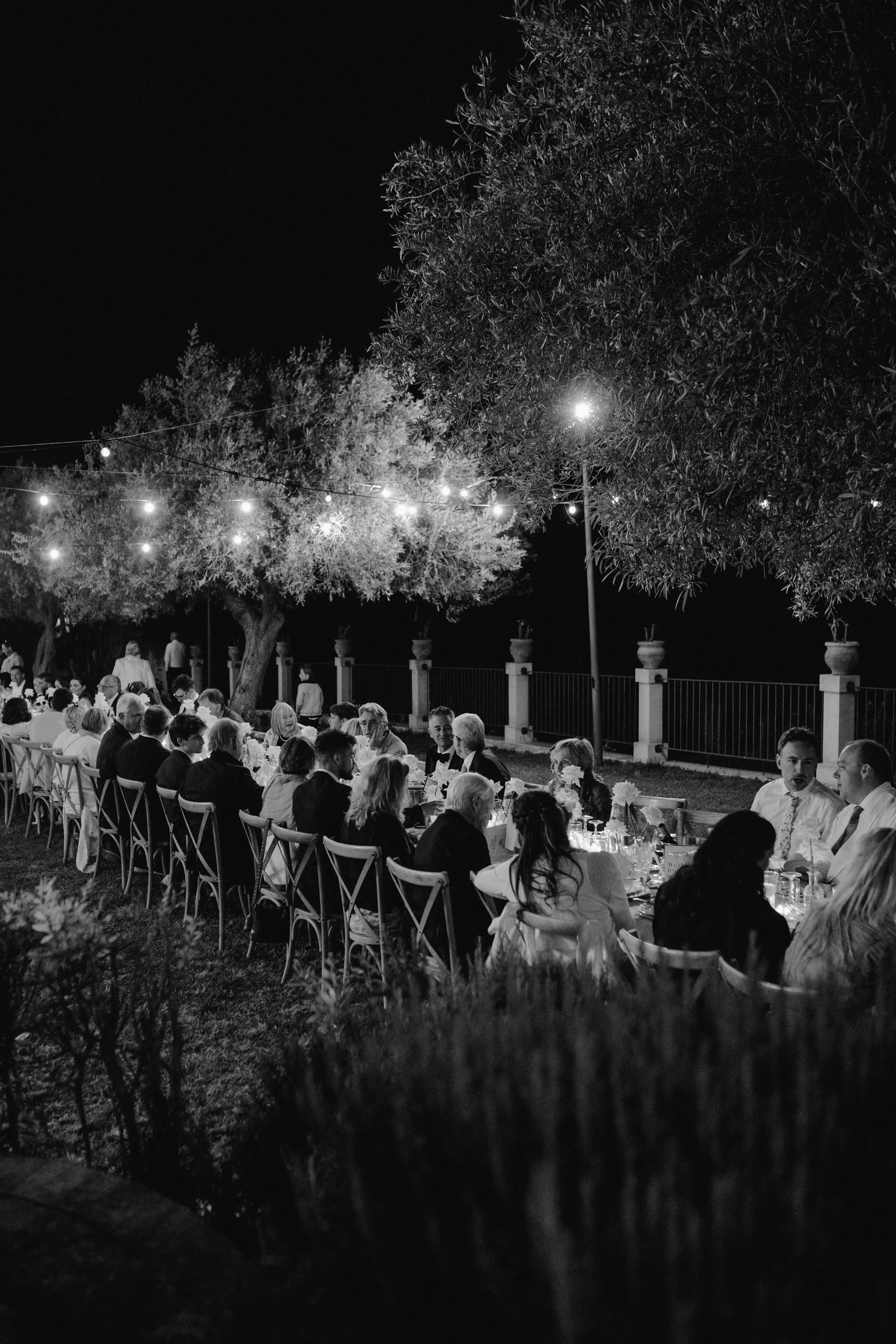 Black and white nighttime wedding reception with guests dining under string lights and olive trees in Sicily.